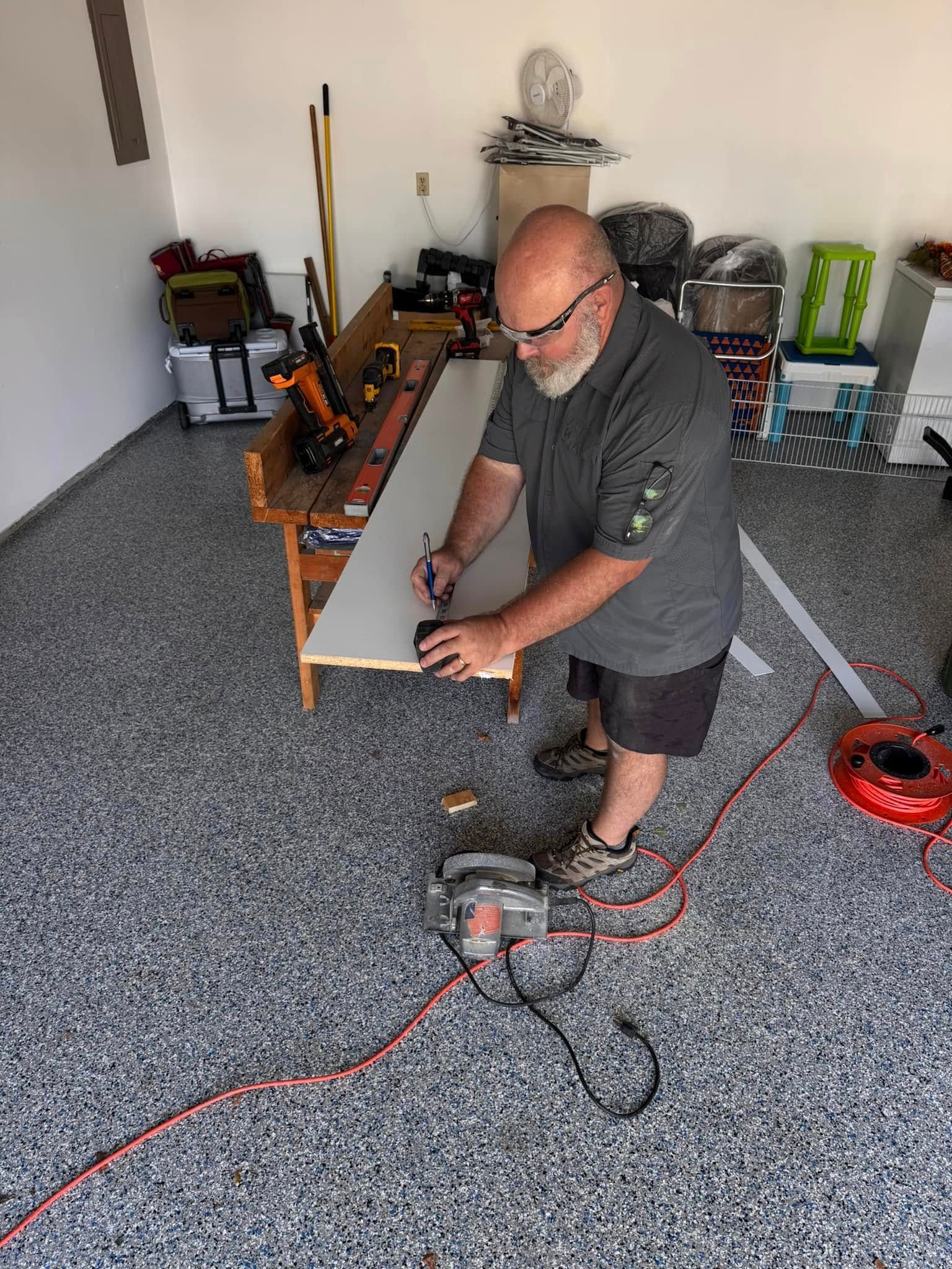 A person marks a measurement on a wooden board placed on a workbench in a garage, with a circular saw on the floor nearby.