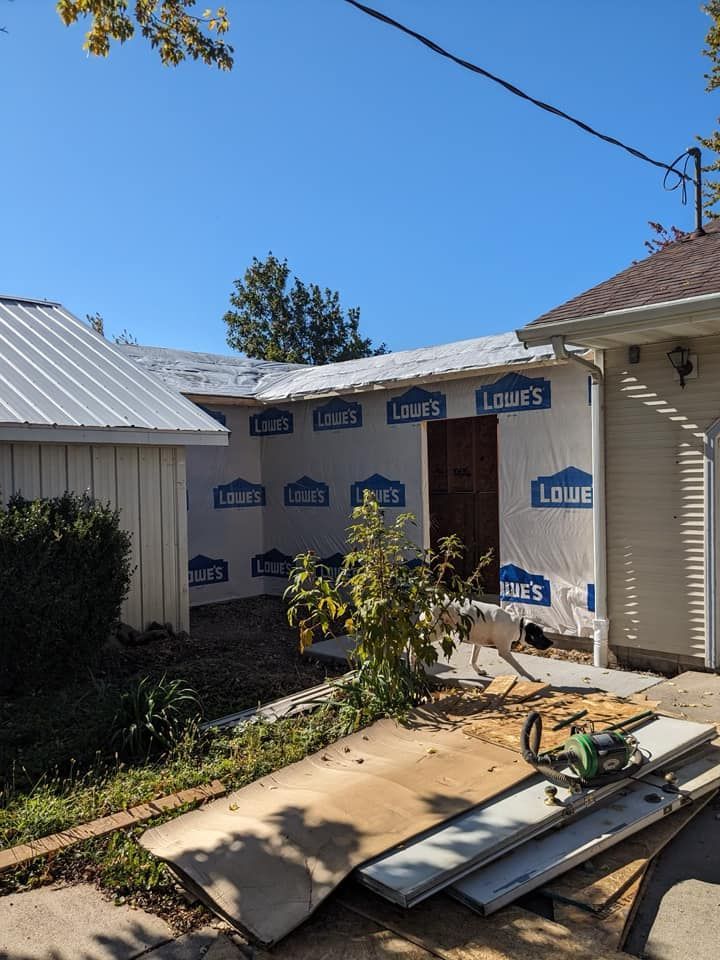A house exterior under construction, with Lowe's house wrap on the walls, a metal roof, and building materials on the ground.