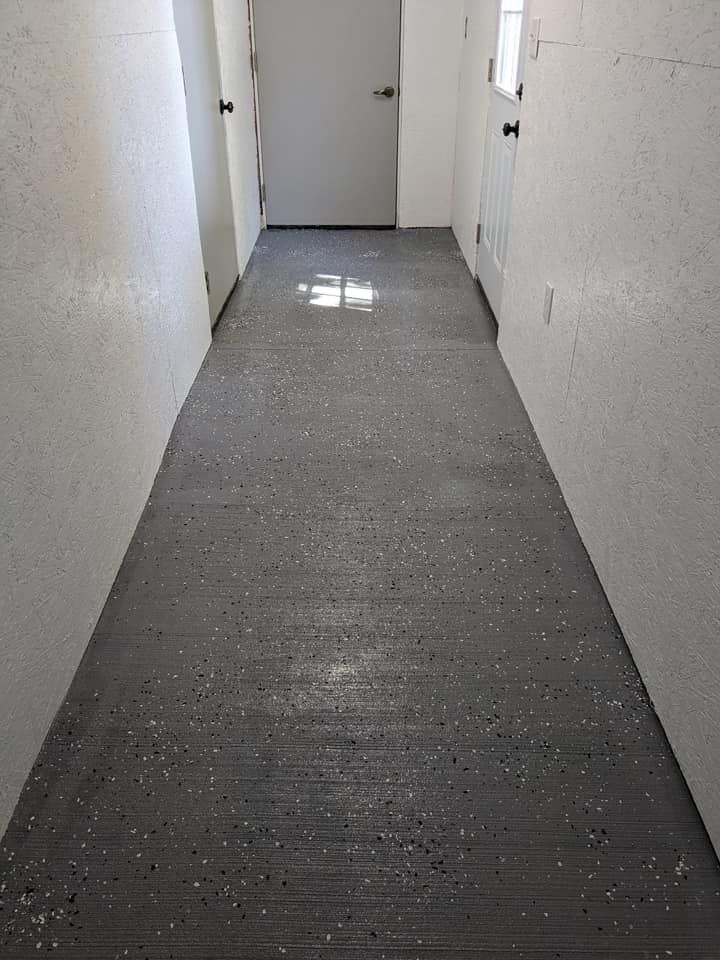 A narrow hallway with white walls and a gray epoxy floor with white speckles, leading to a closed door at the end.