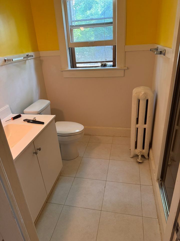 A small bathroom with white cabinets, a toilet, a tall white radiator, and yellow walls over a white wainscot.