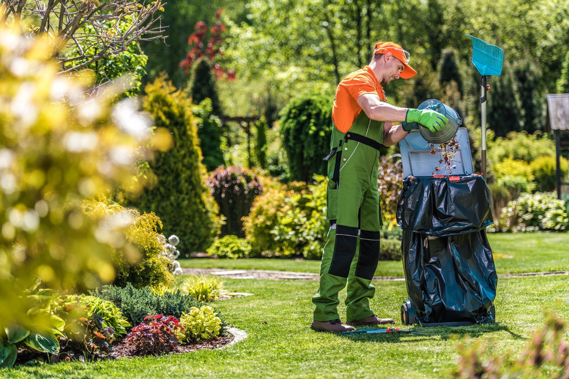 Man in green overalls and orange shirt emptying yard waste into a large black bag in a sunny garden.
