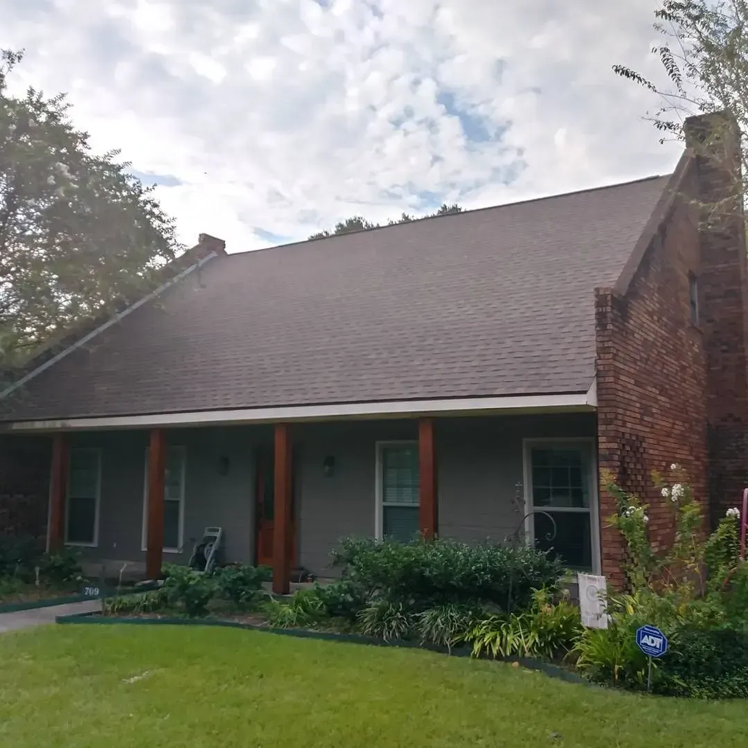 House with gray siding, brown roof, brick chimney, and porch with brown support beams. Green lawn and bushes.