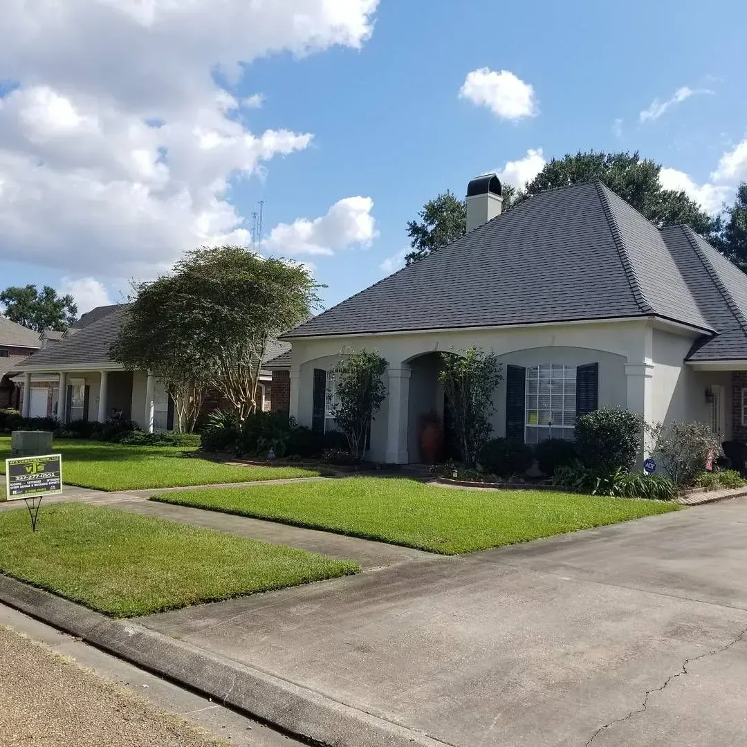 A light-colored house with a gray roof on a sunny day. Green lawn and trees in front.