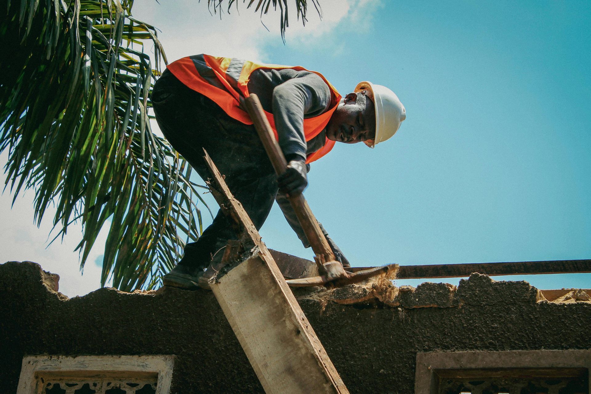 Construction worker repairs a roof, wearing a hard hat and orange vest, against a blue sky.