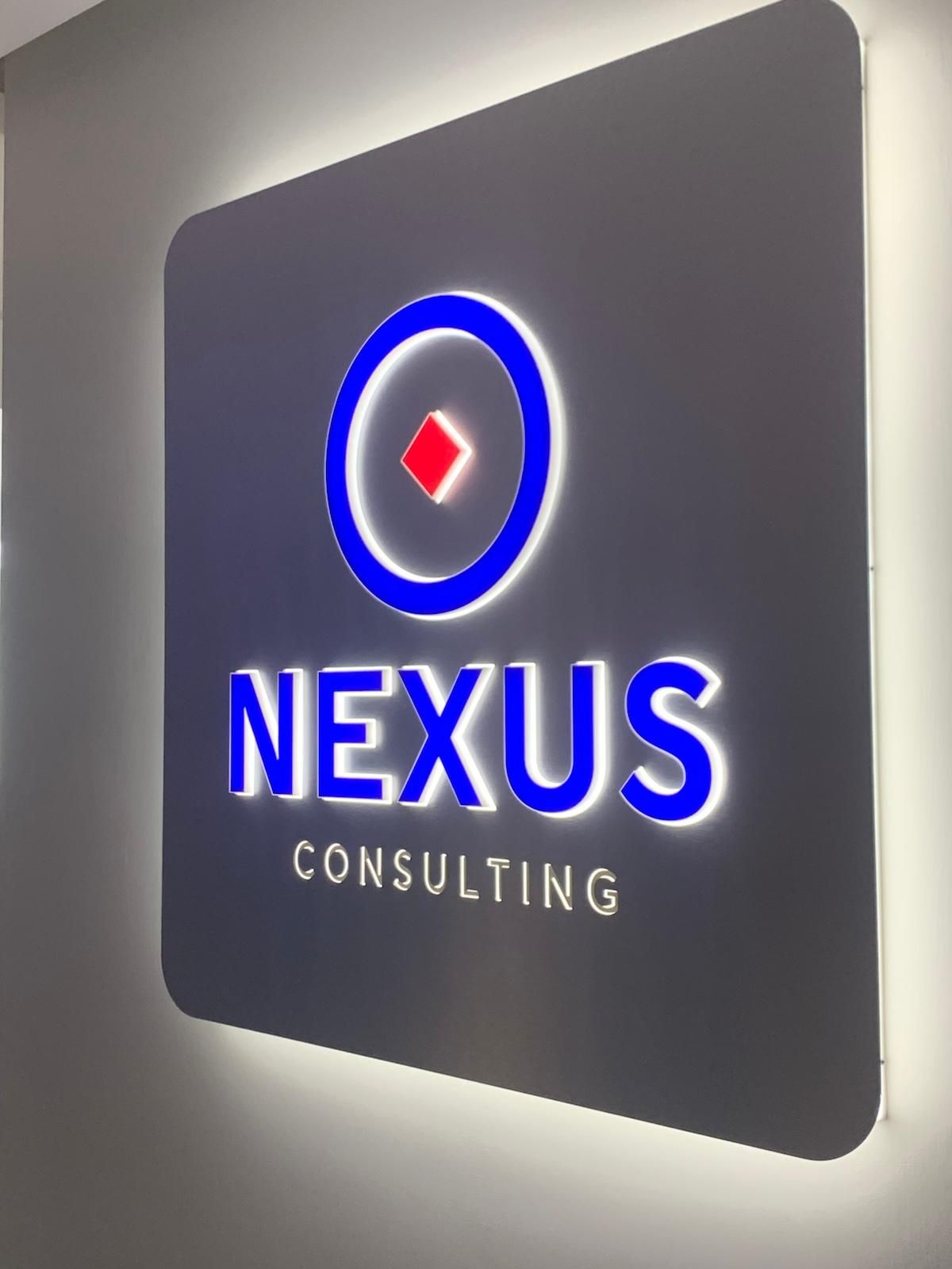 Insegna nera con angoli arrotondati, scritta "NEXUS CONSULTING" in bianco, cerchio blu con diamante rosso.