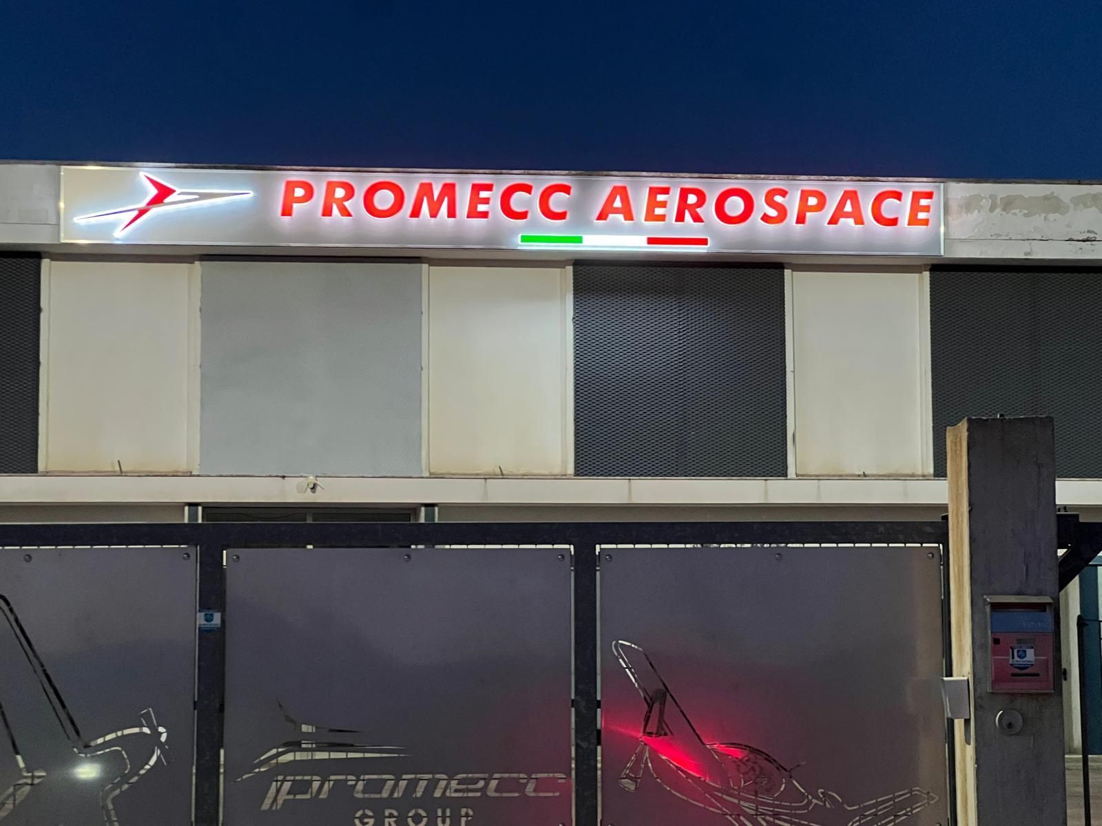 Insegna del Promecc Aerospace Group, con logo e colori della bandiera italiana. Illuminata di notte.