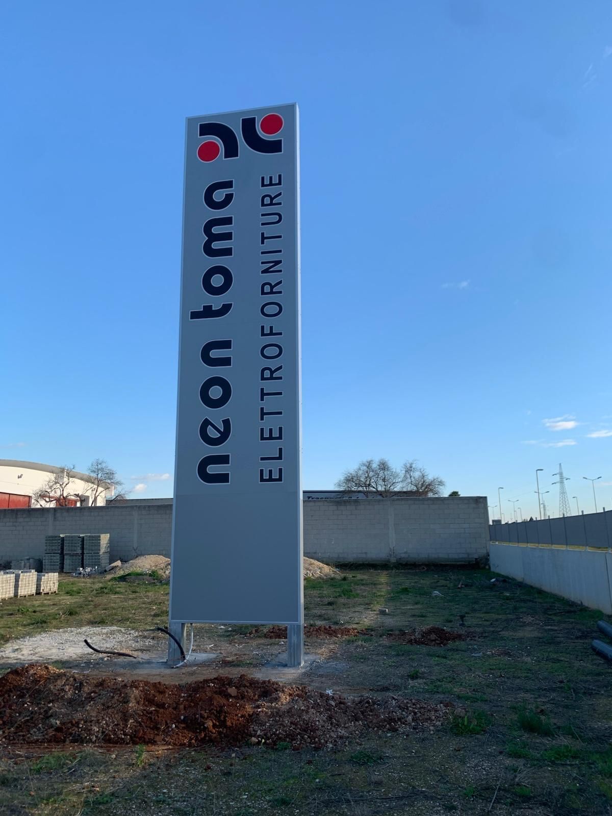 Insegna per Neon Toma Elettroforniture, alta, grigia, logo rosso, contro il cielo azzurro.