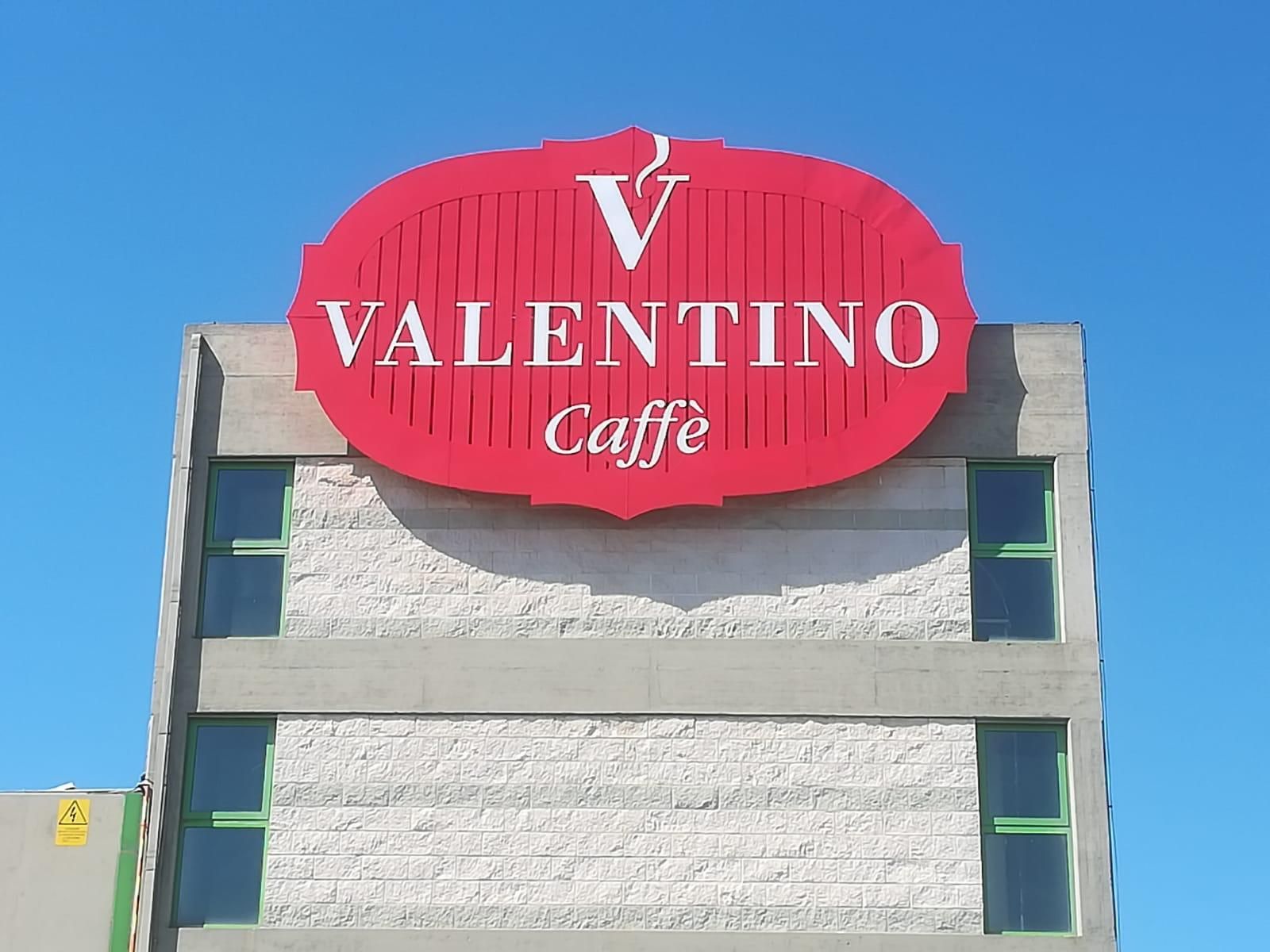 Insegna del Valentino Caffè in cima a un edificio grigio chiaro con cielo azzurro. Logo rosso con testo bianco.