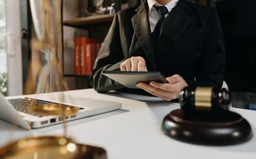 Lawyer in black suit uses tablet at desk with laptop, gavel, and books.