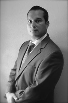 Man in suit and tie, looking serious. Leaning against a wall, arms crossed. Black and white photo.