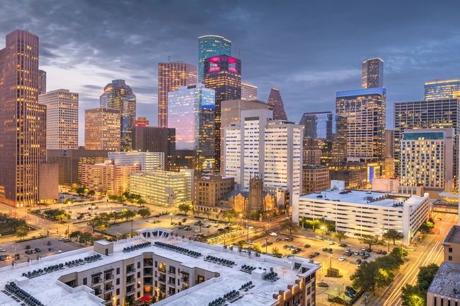 Houston skyline with a river and lush green trees under a blue sky with fluffy clouds.
