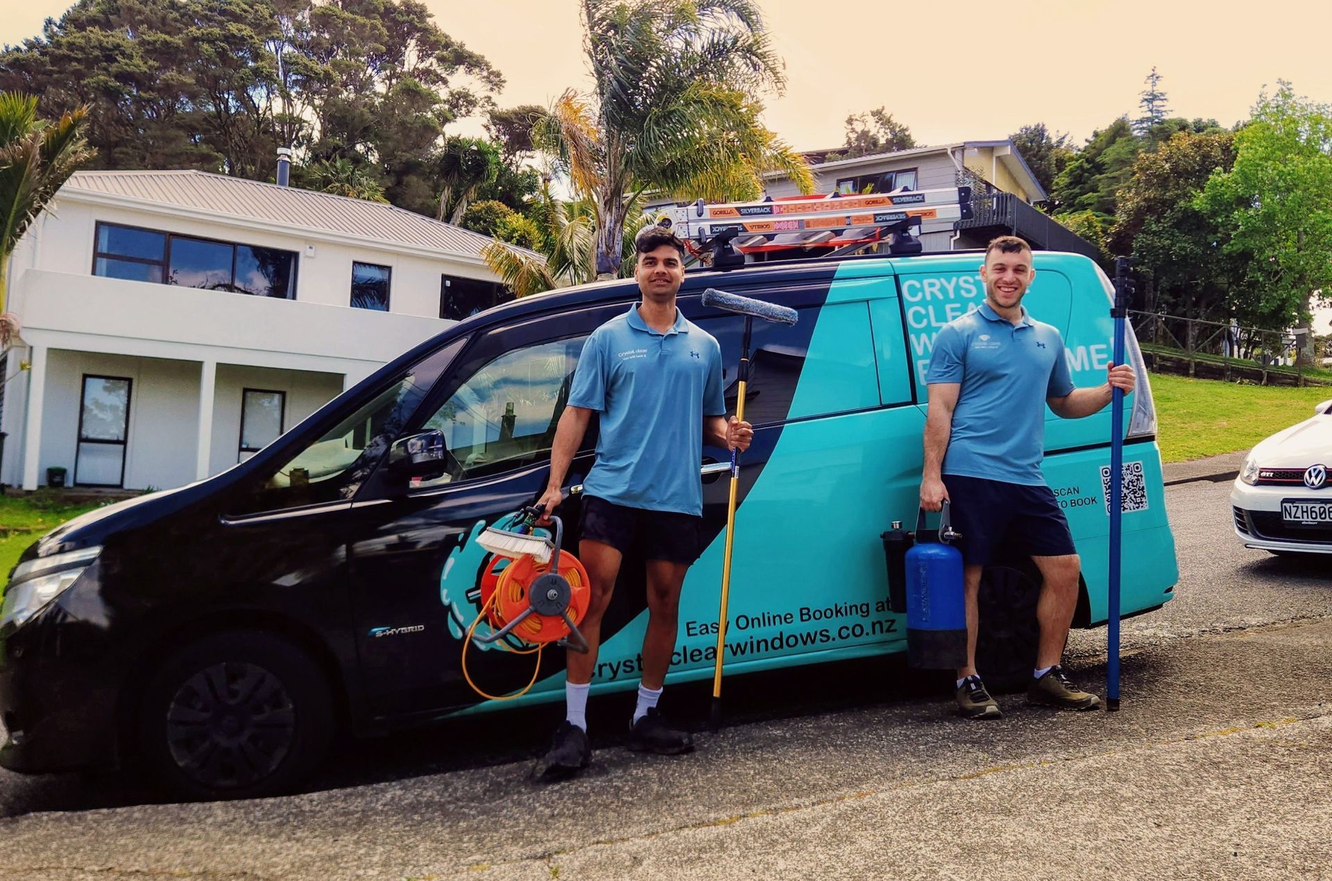 Two men in matching shirts stand by a cleaning van, holding equipment. Black van with teal logo in residential setting.