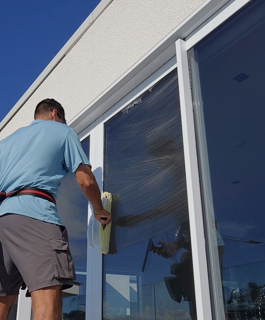 Man in hat cleans tall windows with a long-handled squeegee on a sunny day.