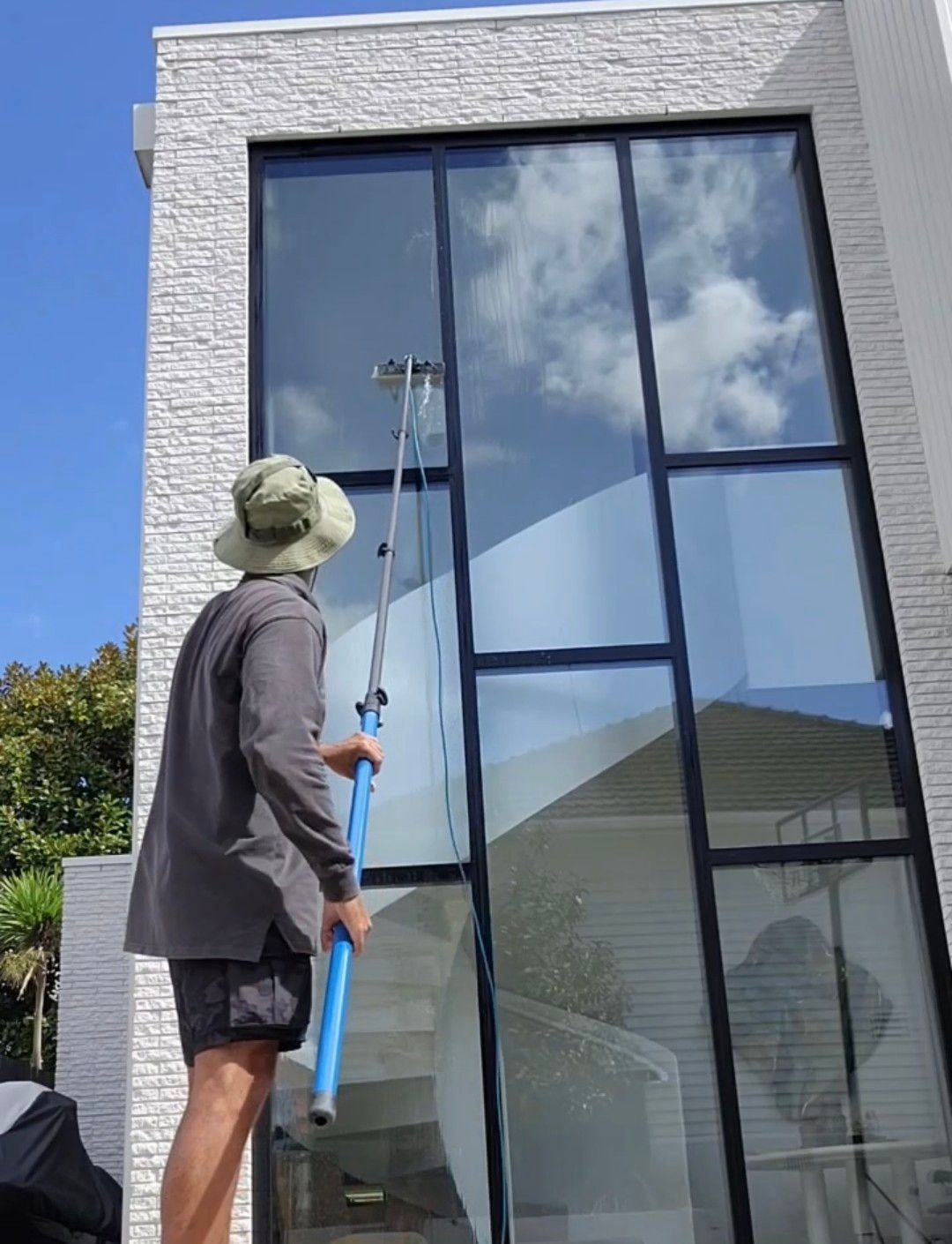 A man in shorts and a hat cleans a tall window with a long-handled squeegee, white building, blue sky.