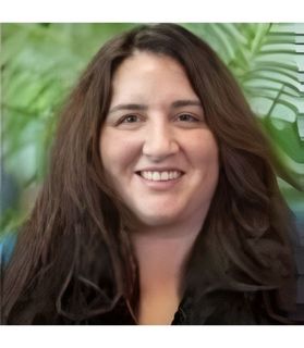 Woman with long brown hair smiling, in front of a green leafy background.