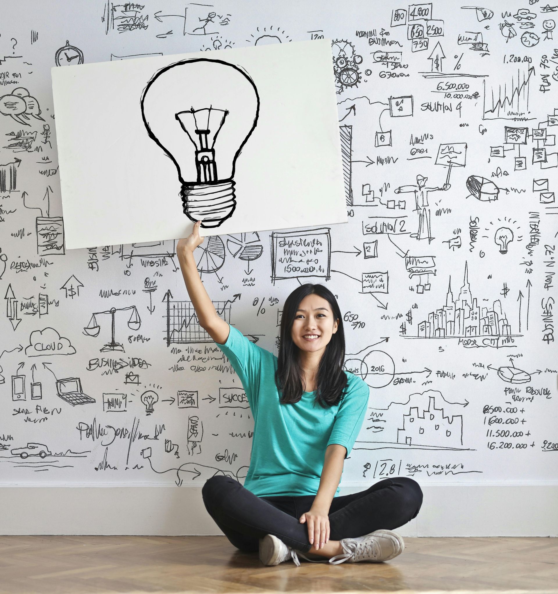 A woman is sitting on the floor holding up a sign with a light bulb drawn on it