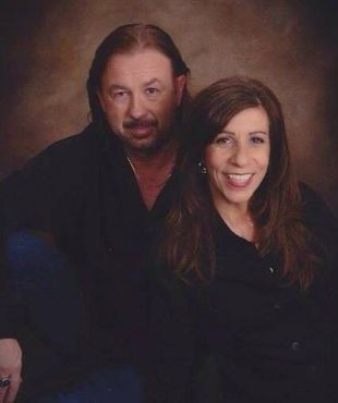 A middle-aged couple poses in a studio portrait. The man has long hair and a mustache. The woman smiles, and both wear black shirts.
