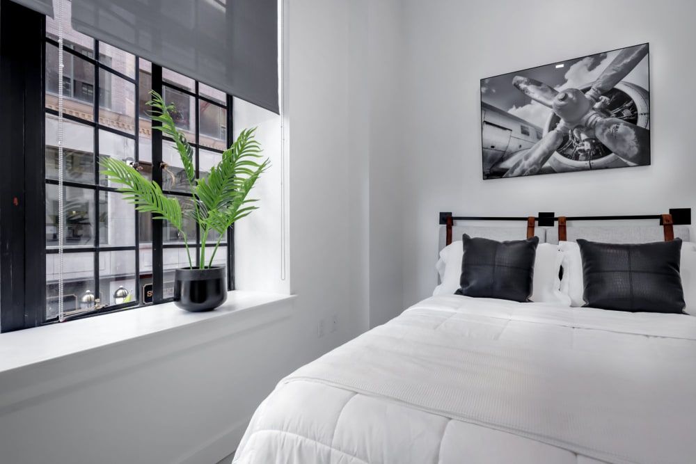 A bedroom with a bed , window , plant and painting on the wall.