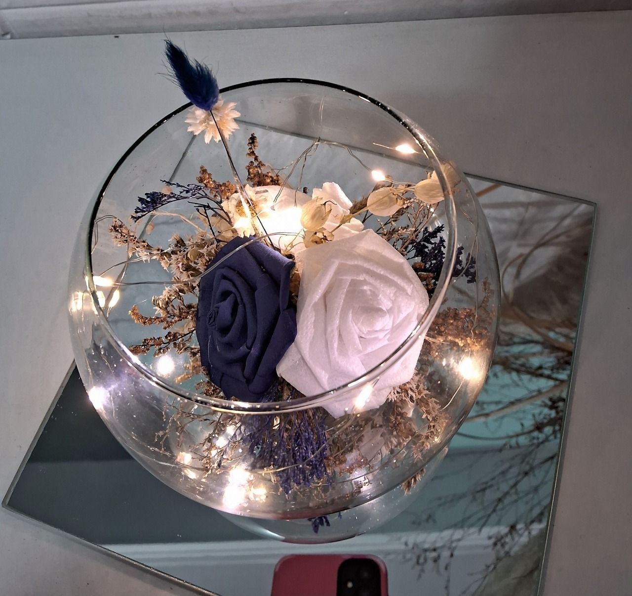 Un terrario de cristal con flores de tela azules y blancas, luces de hadas y follaje seco, sobre una superficie espejada.