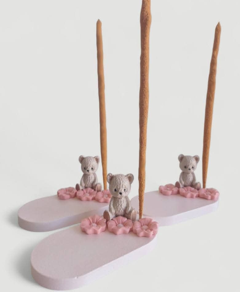 Tres porta inciensos rosas, cada uno con un osito de peluche, flores rosas y una varilla de incienso marrón.