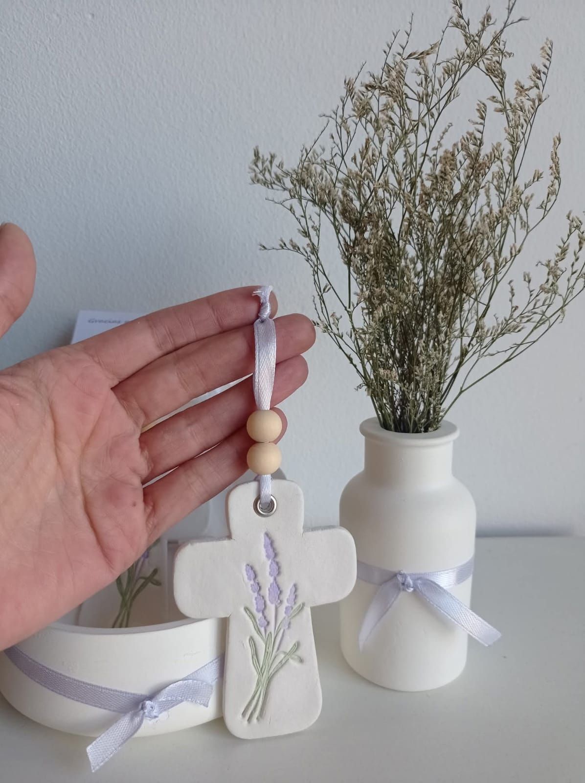 Mano sosteniendo un adorno de cruz blanca con diseño de lavanda, junto a un jarrón con flores secas.