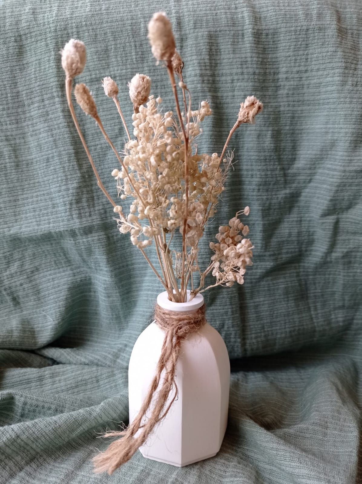 Jarrón blanco con flores secas y detalles de cordel, sobre un fondo de tela verde drapeada.