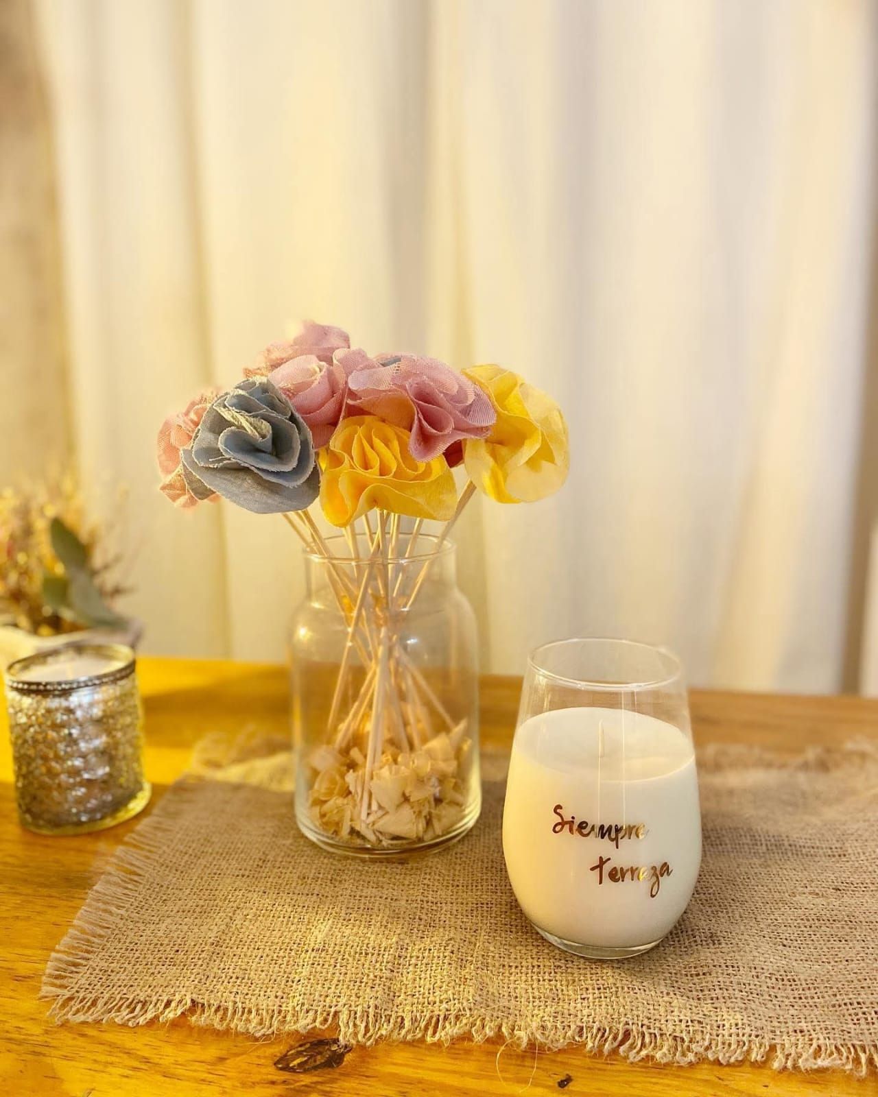 Ramo de flores en tonos pastel en un tarro de cristal, con una vela encendida al lado sobre una mesa de madera.