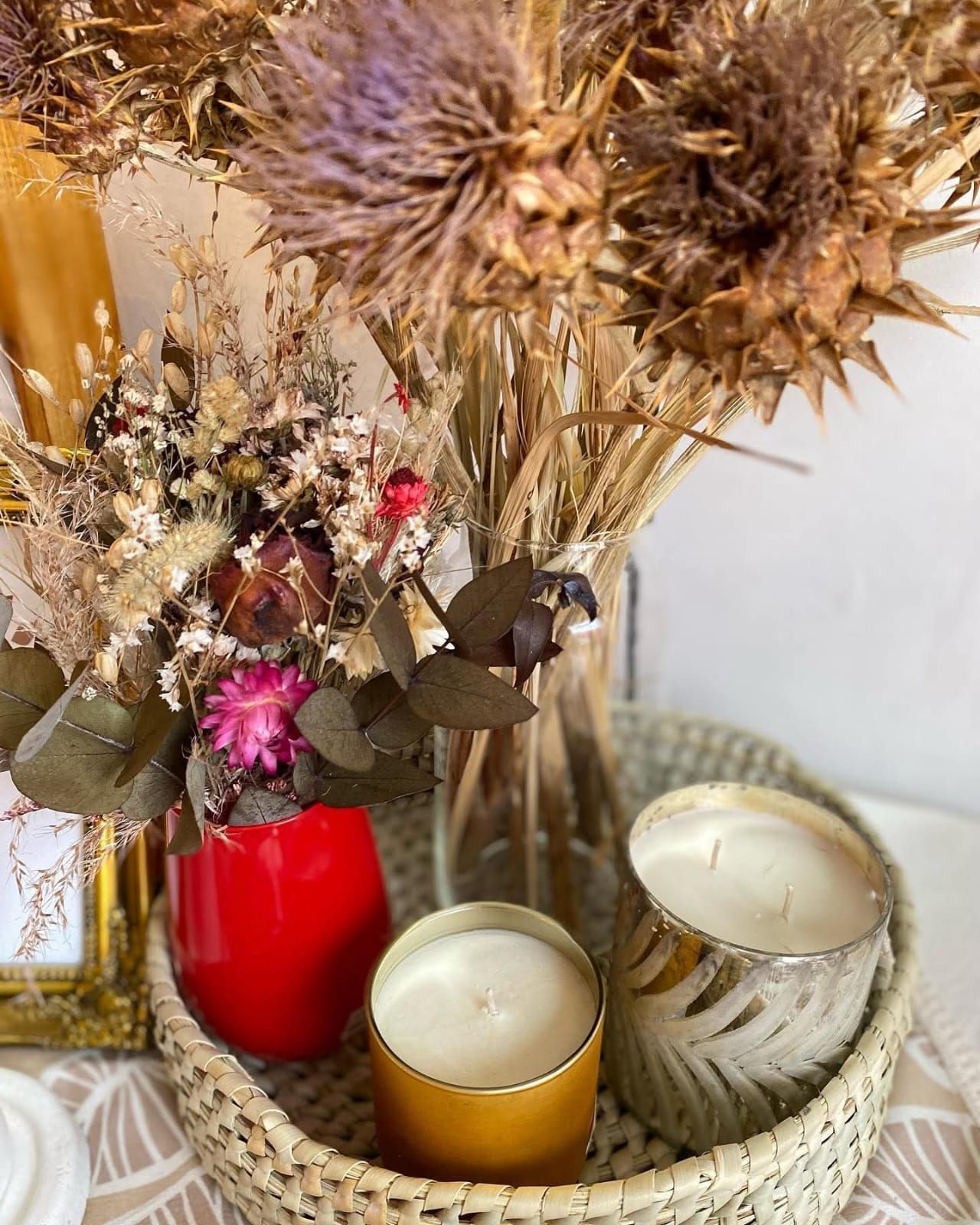 Flores secas y velas en una bandeja de mimbre. Un jarrón rojo contiene flores. Candelabros dorados y plateados.
