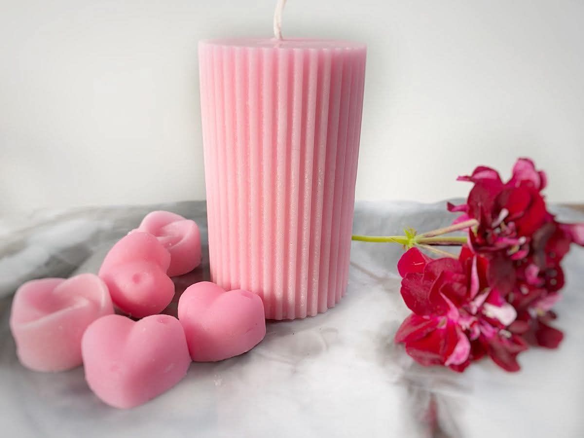 Vela rosa acanalada con cera derretida en forma de corazón y flores rojas sobre una superficie de mármol.