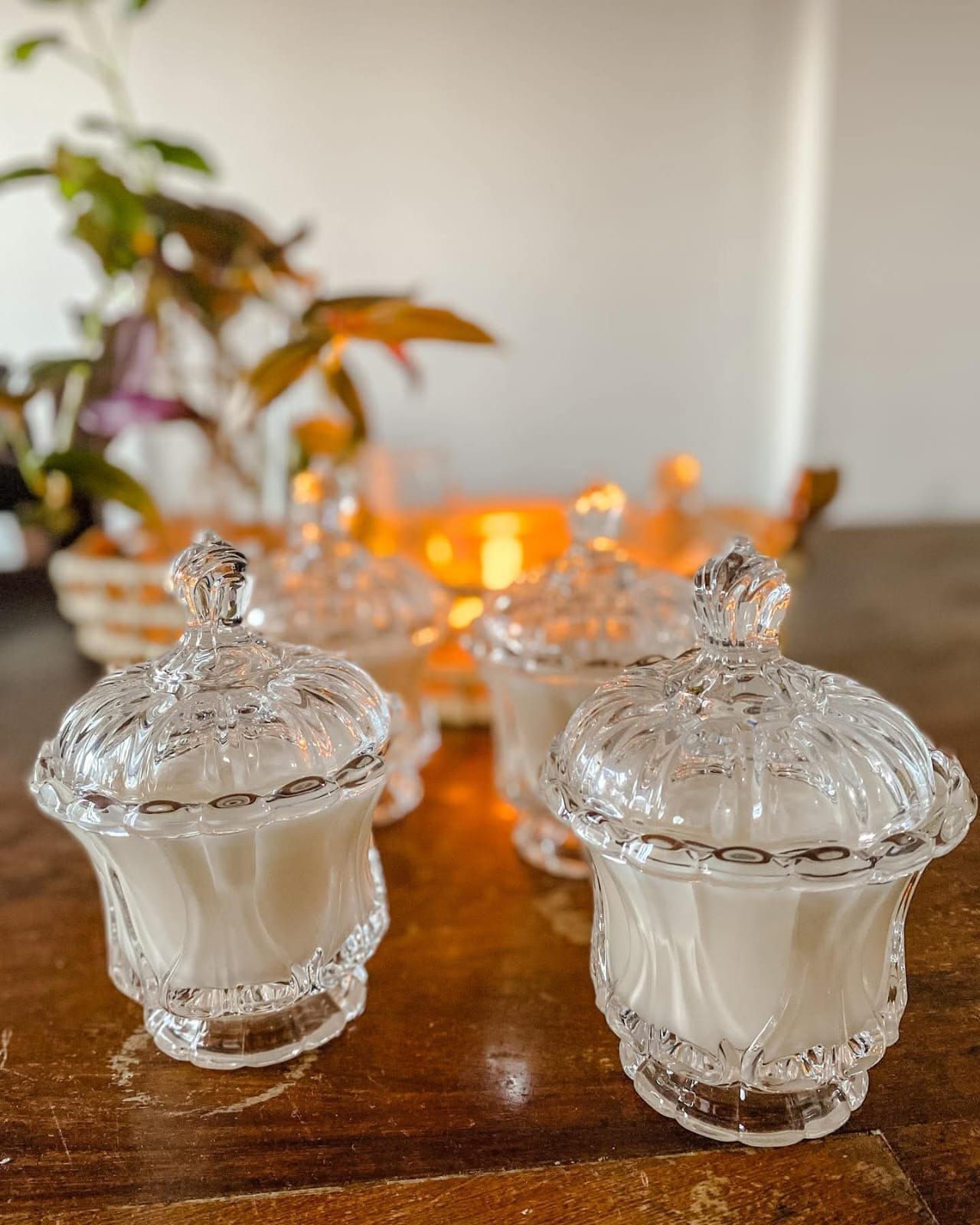 Cuatro frascos de vidrio ornamentados con tapa, que contienen velas blancas, sobre una mesa de madera.