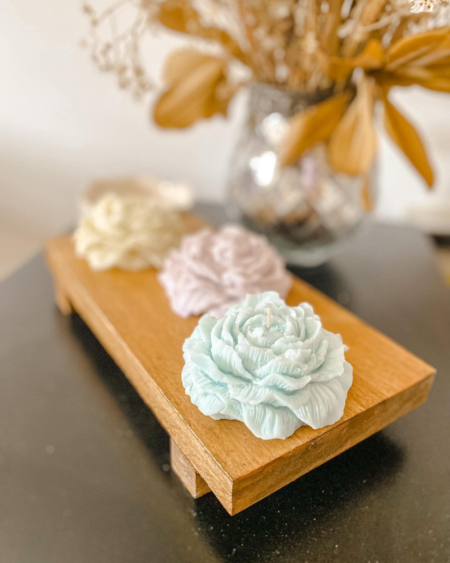 Tres velas en forma de flor de colores pastel sobre una bandeja de madera, con flores secas en un jarrón al fondo.