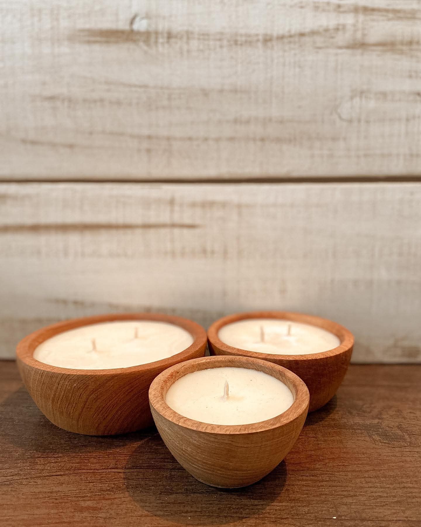 Tres velas de cera de soja en cuencos de madera sobre una superficie marrón, con un fondo de madera encalada.