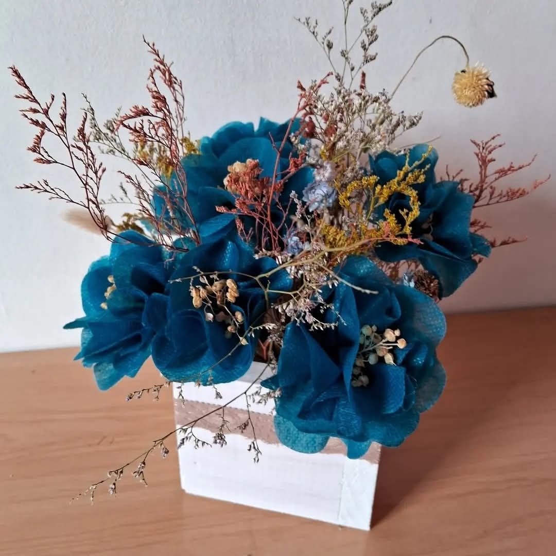 Flores de tela color verde azulado y arreglo floral seco en un jarrón cuadrado a rayas blancas y beige.