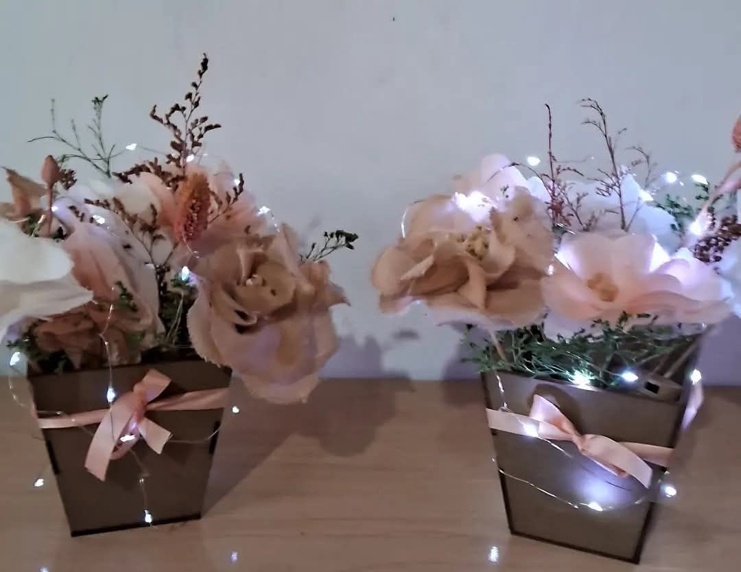 Dos arreglos florales en macetas cuadradas marrones con cinta rosa y luces de hadas.