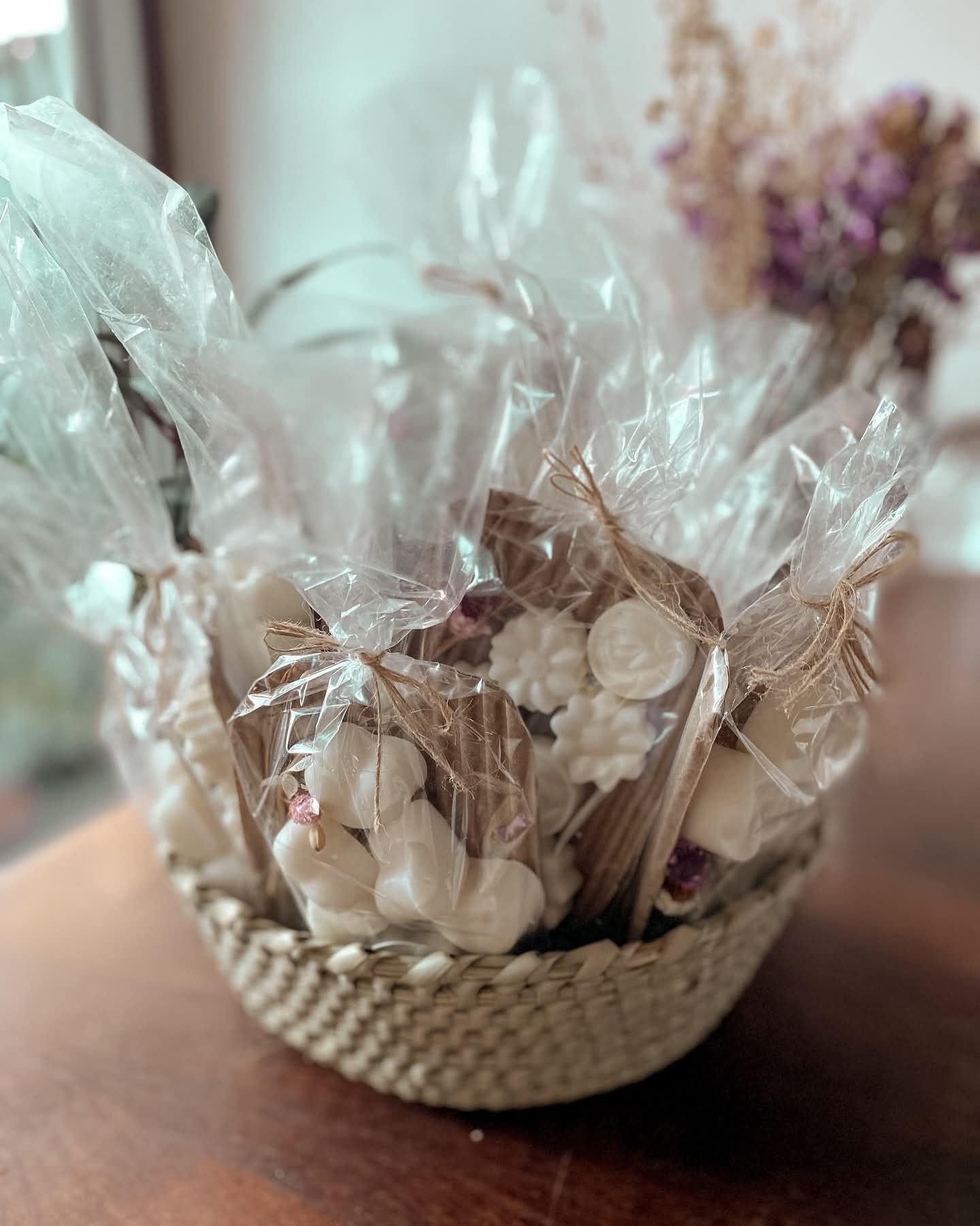 Sobre una mesa de madera, una cesta de mimbre llena de jabones blancos envueltos y atados con rafia.