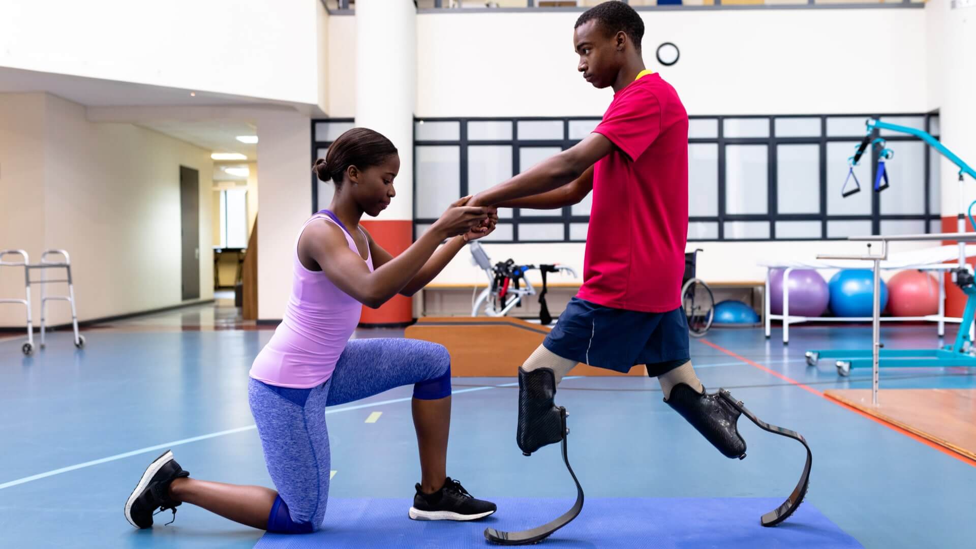 Side view of African-american physiotherapist helping disabled African-american man walk with prosthetic leg in sports center. Sports Rehab Centre with physiotherapists and patients working together