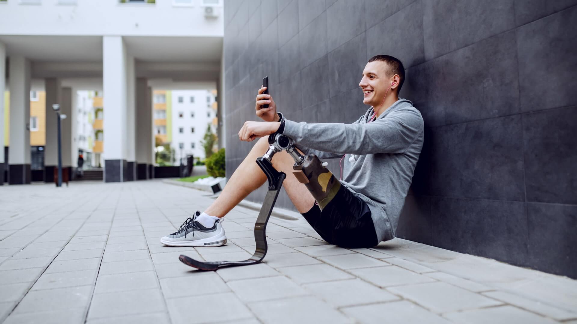Young smiling positive handsome caucasian sportsman with artificial leg sitting on ground, leaning on the wall, listening music and texting on smart phone.