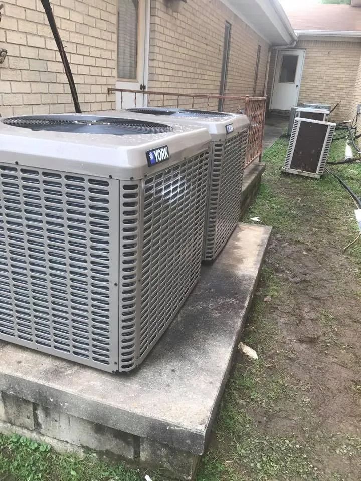 Two outdoor HVAC units on a concrete pad beside a brick house with a grassy yard.