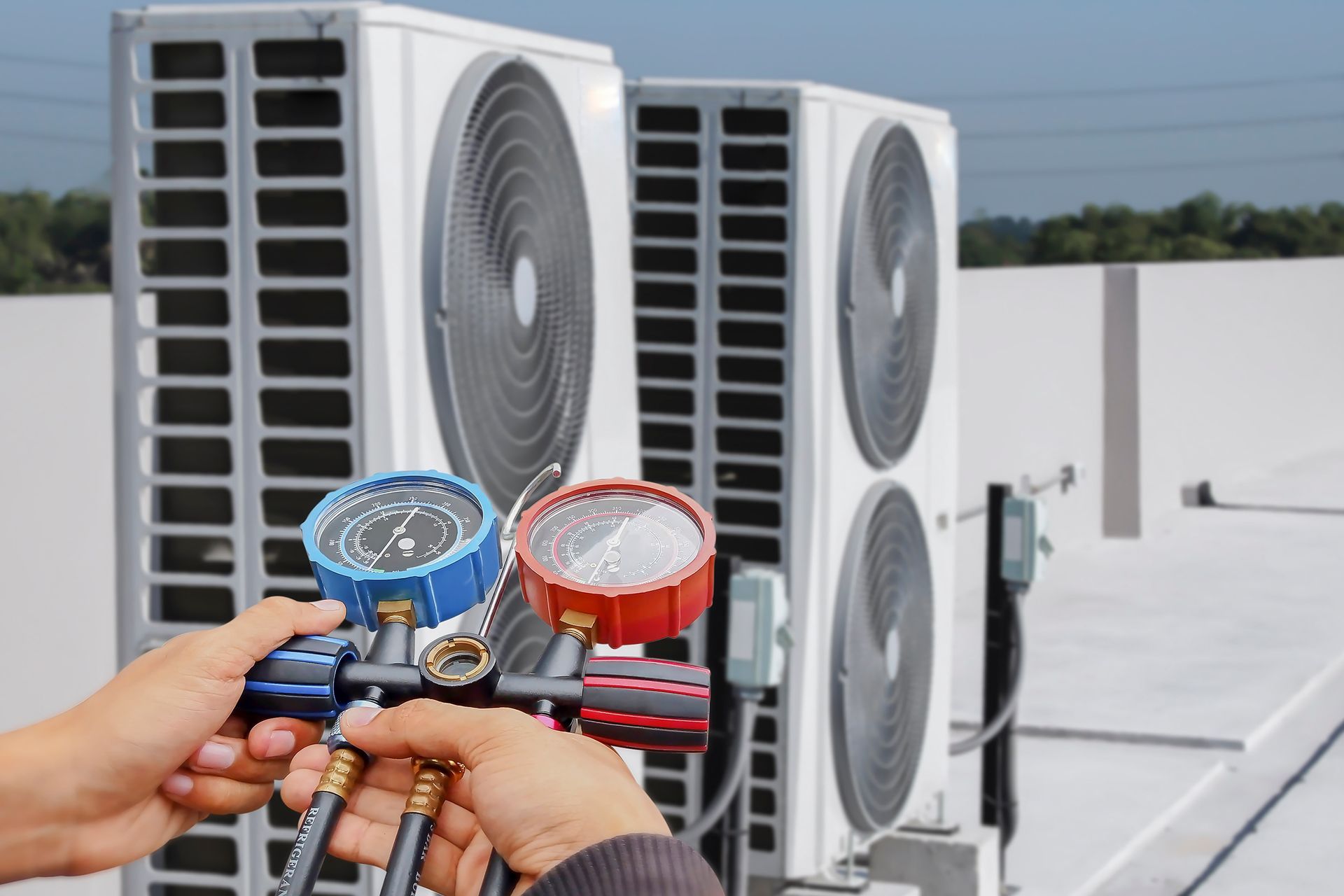 Hands holding HVAC manifold gauges in front of outdoor air conditioning units on a rooftop.
