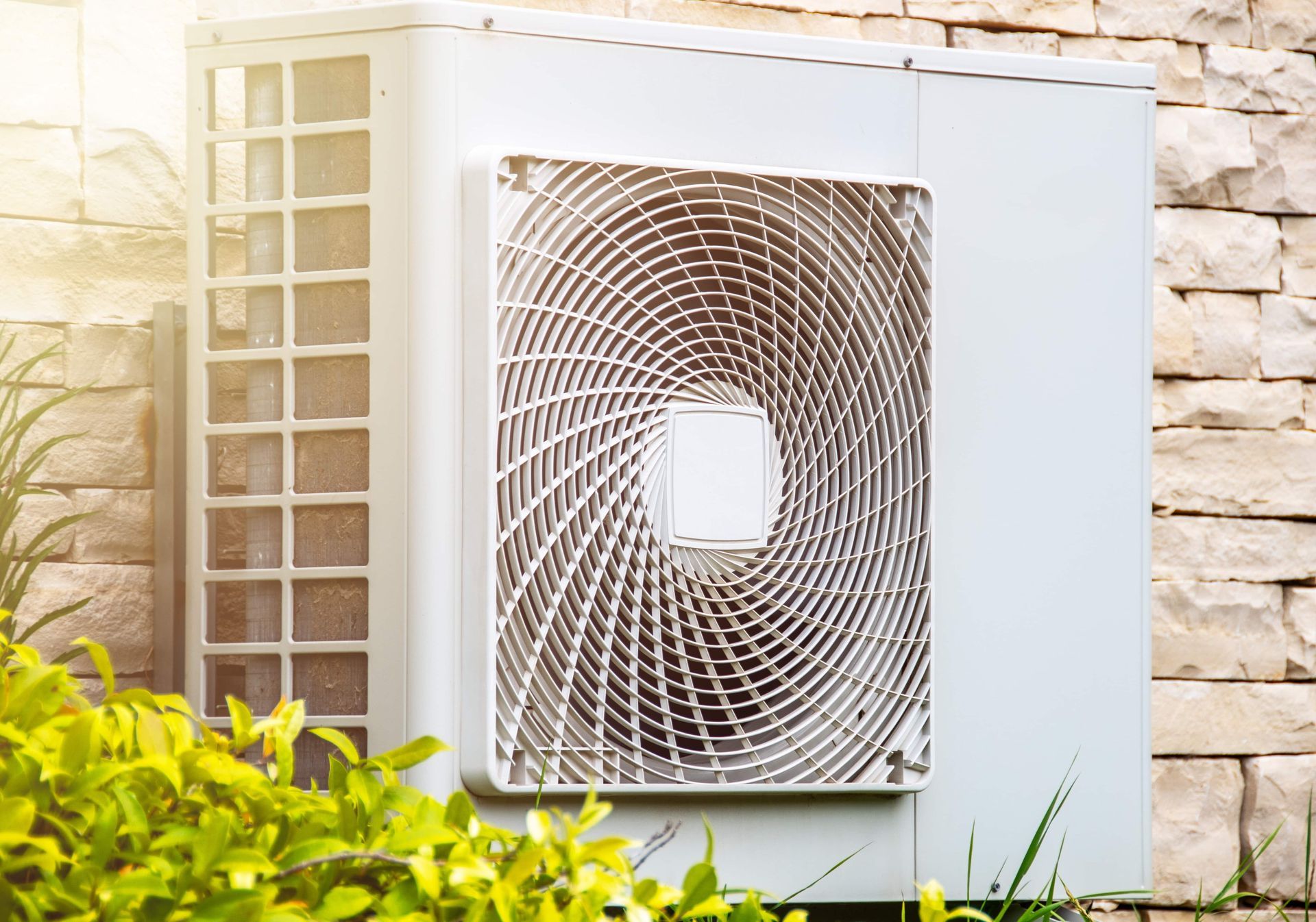 Outdoor air conditioner unit mounted on a stone wall beside a window, partly framed by green shrubs