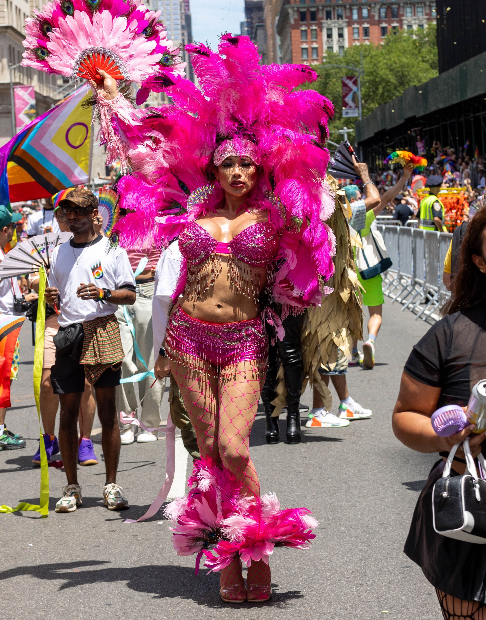 Person in pink feathered costume at a parade, with a headdress, holding a fan.