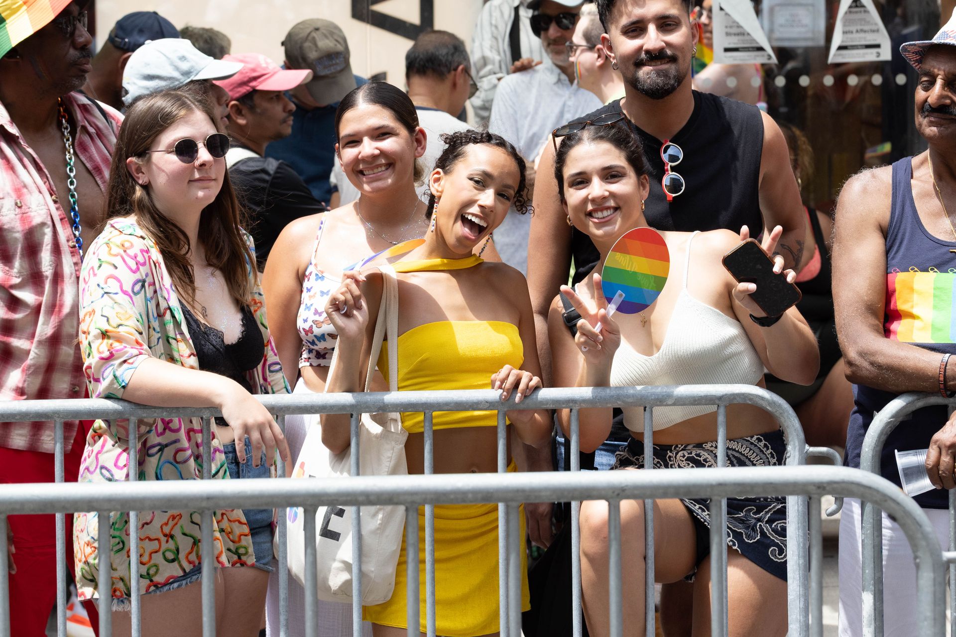 People at a Pride parade, smiling and waving. Rainbow flag and fence in front of them.