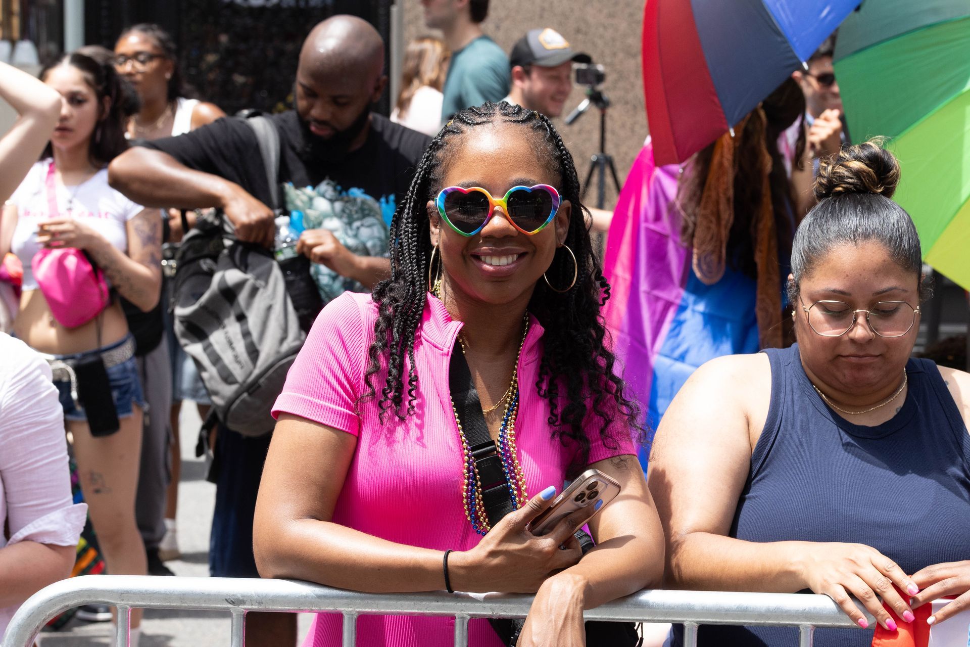Woman with heart-shaped rainbow sunglasses smiles at a Pride parade; people, rainbow flag, and fence are in the background.