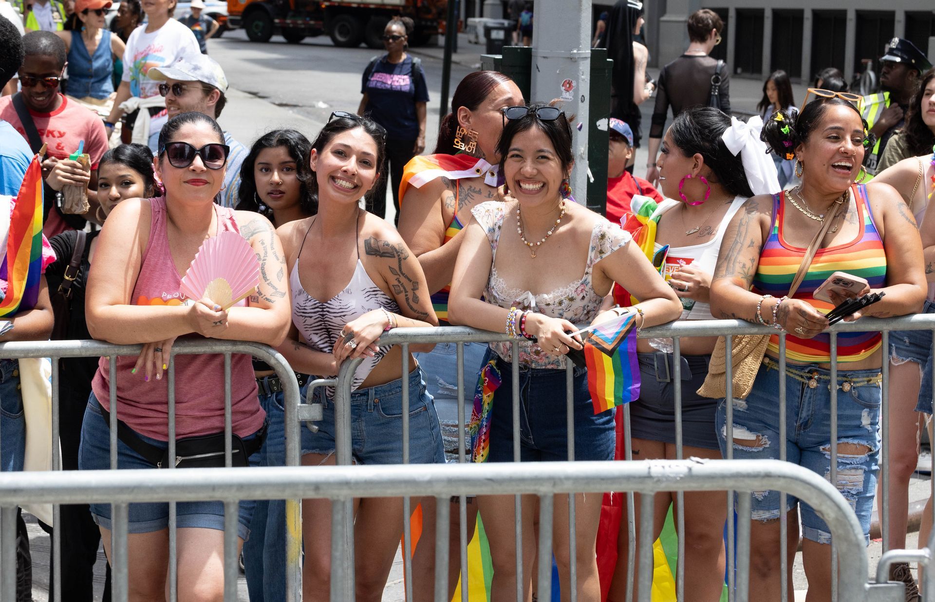 People watching a Pride parade, leaning on a barricade, smiling and holding rainbow flags in a sunny outdoor setting.