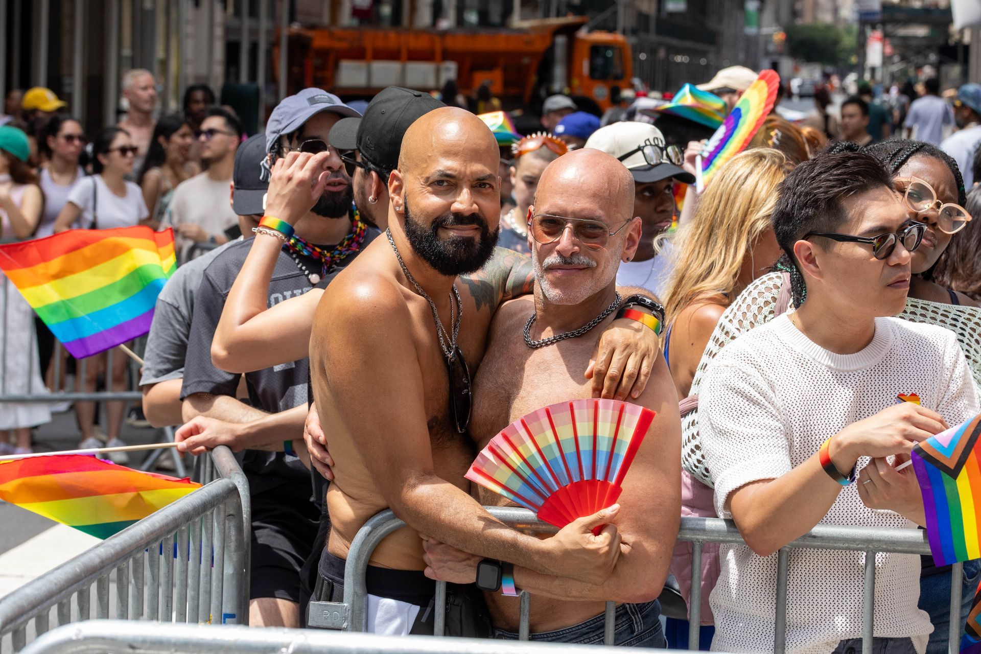 Two men embrace at a Pride parade, one holding a rainbow fan. Many people and rainbow flags are visible.