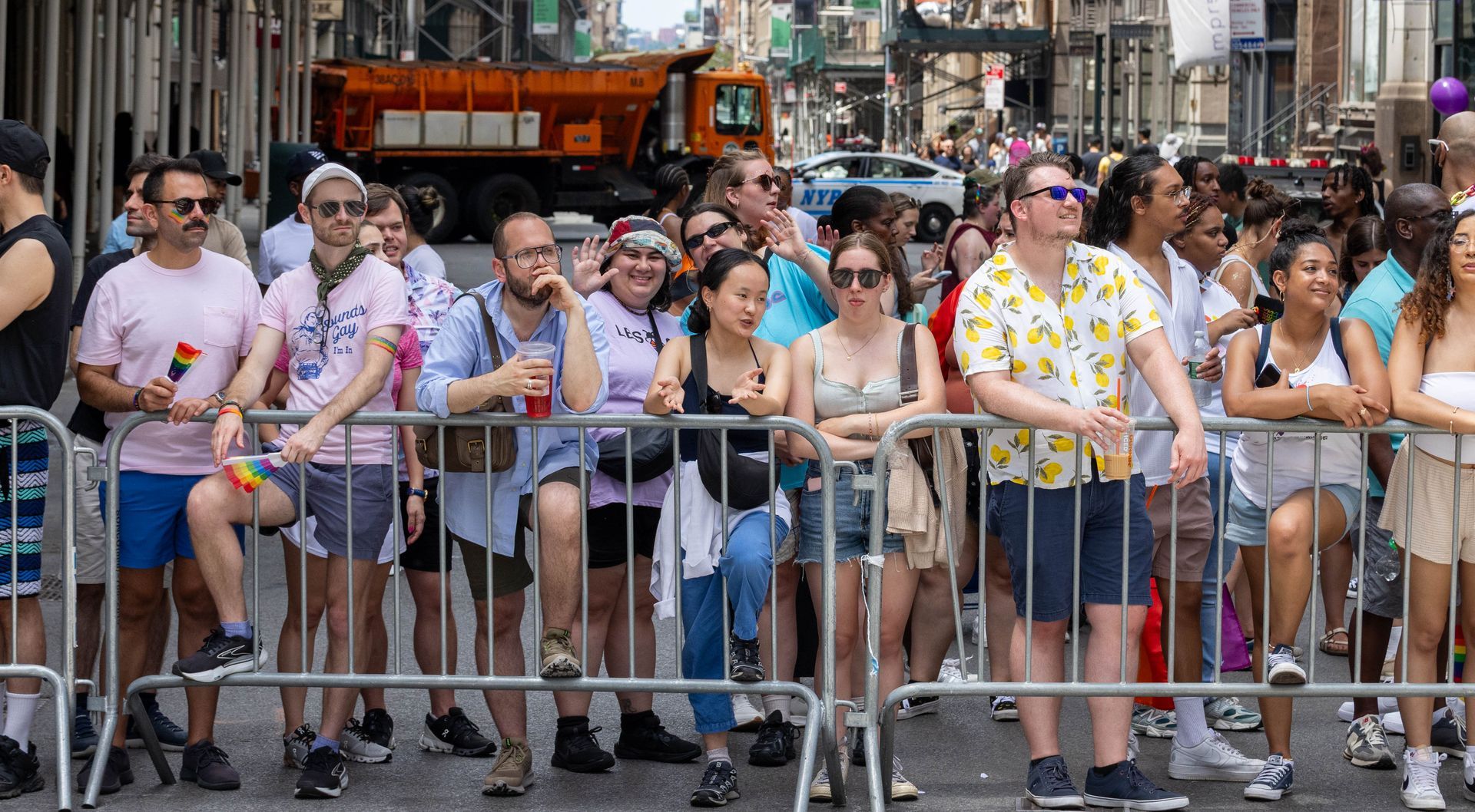 Crowd watches a Pride parade from behind a metal barrier. People of diverse ages and appearances. Street scene with a truck in the background.