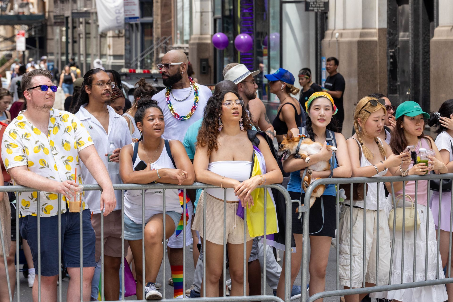 People watching a parade, some holding onto a metal barrier, outdoors. Various skin tones, fashion, and expressions.