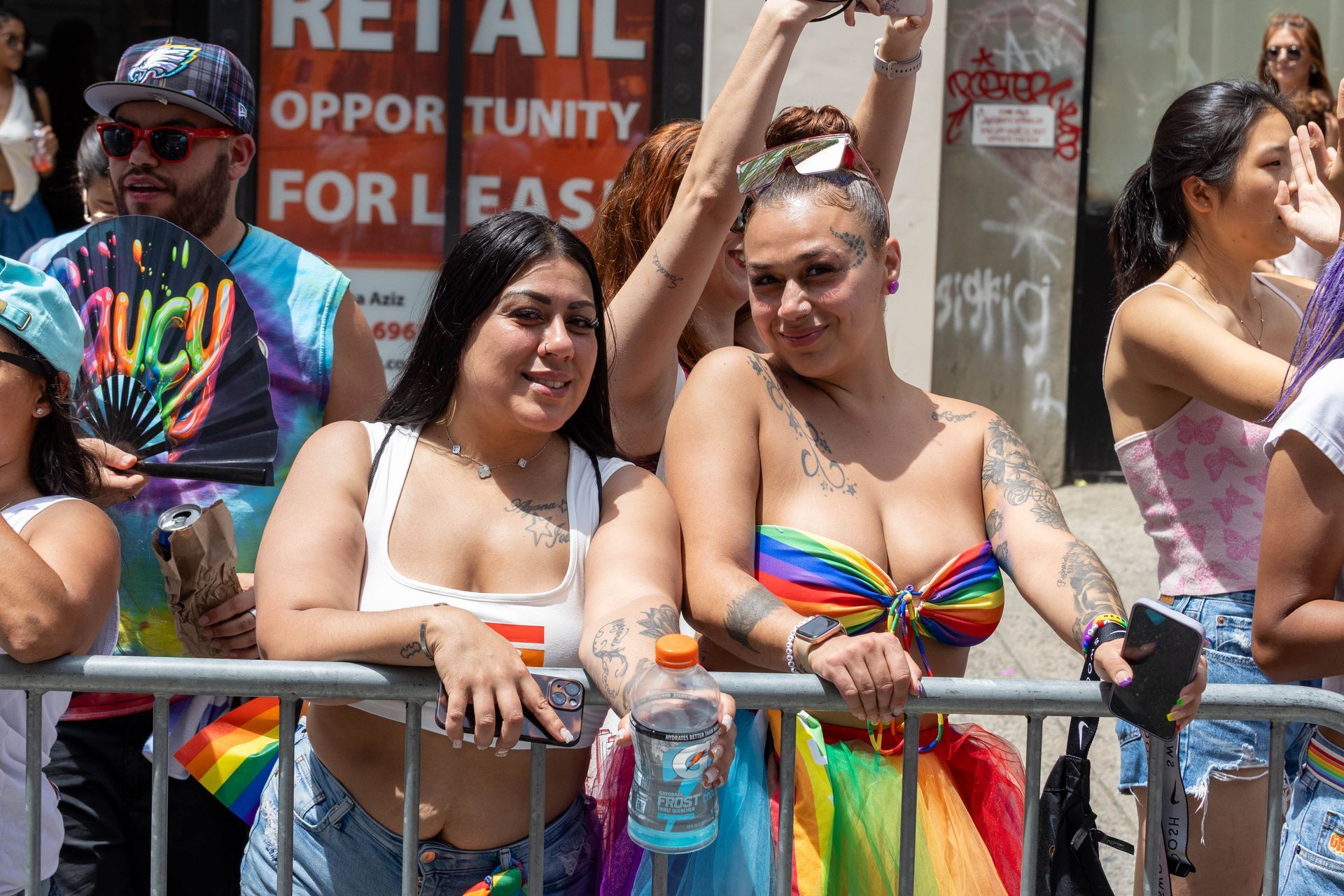 Two women smiling, leaning on a barrier at a Pride parade. One in rainbow top/skirt, the other in white top.