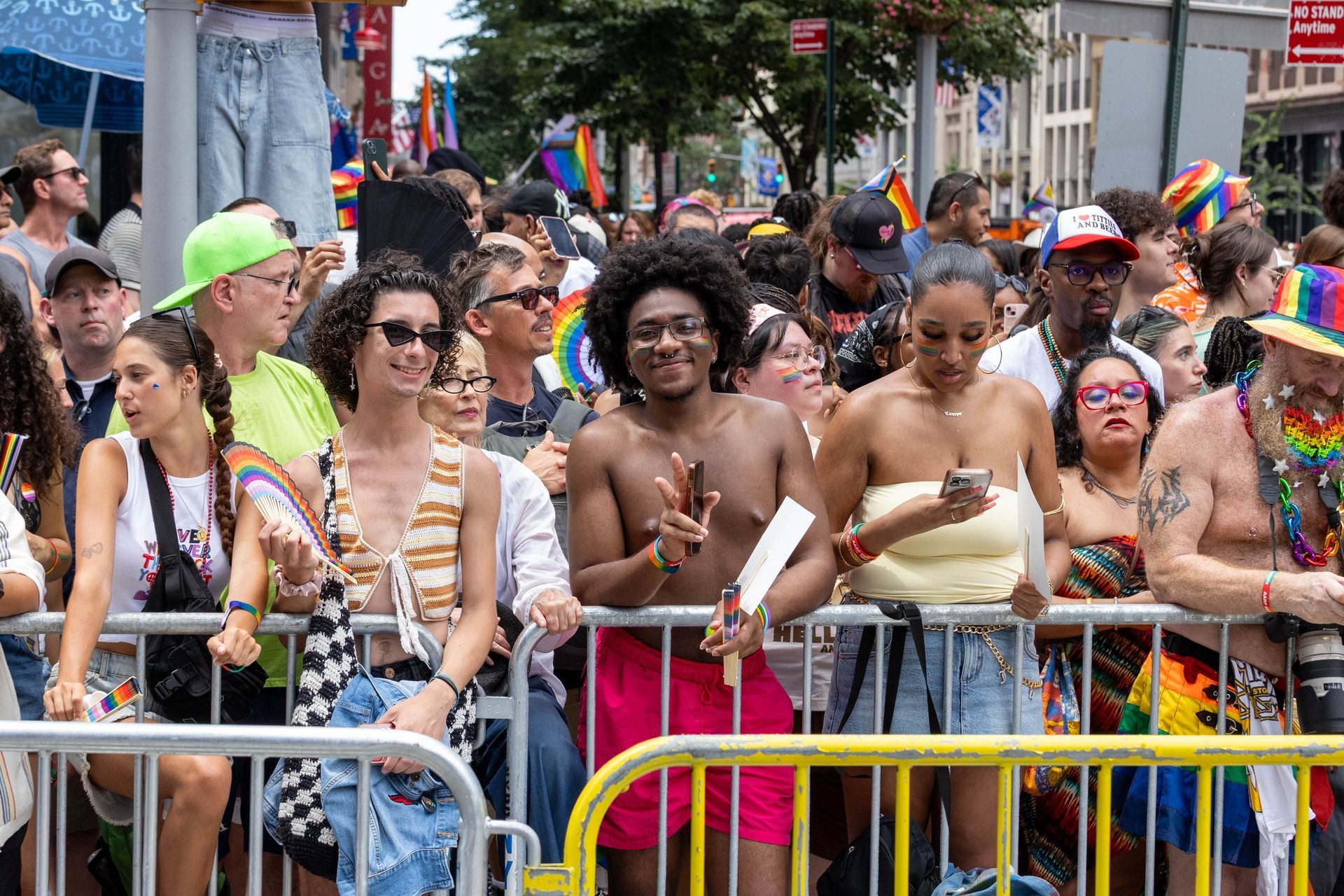People at a Pride parade. Diverse crowd behind a metal barrier. Celebratory expressions and rainbow flags are visible.