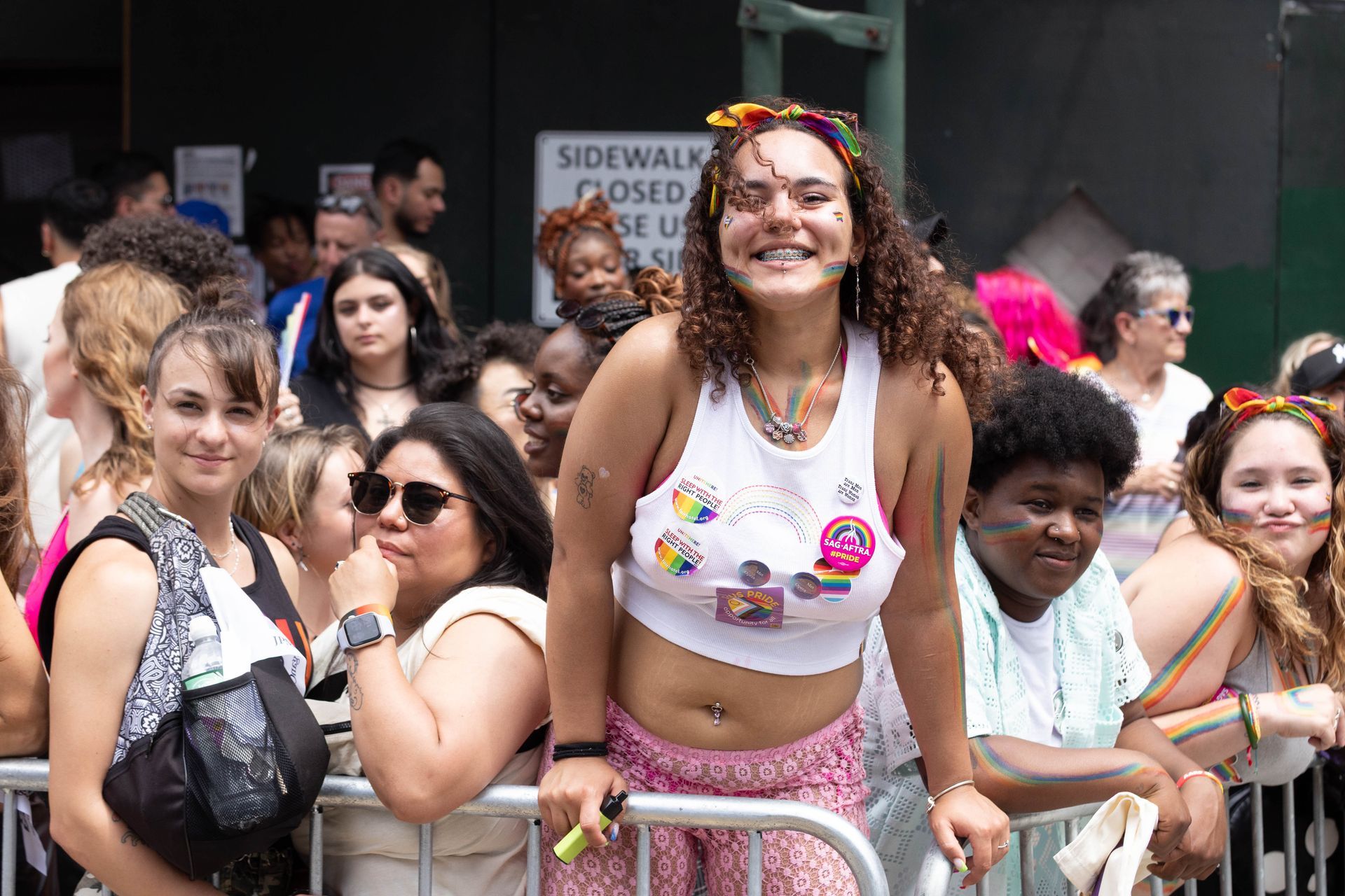 Group of people smiling at a Pride parade, some wearing rainbow colors, standing behind a metal barrier.